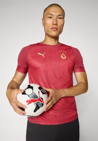 Piros rövid ujjú sportpóló logóval. A játékos egy fekete-fehér futball labdát tart, amelyen piros logó található. Egyszerű háttér.