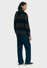 Jeune personne avec des cheveux bouclés portant un pull à capuche rayé marron et noir, un jean bleu foncé et des baskets noires à tige basse.