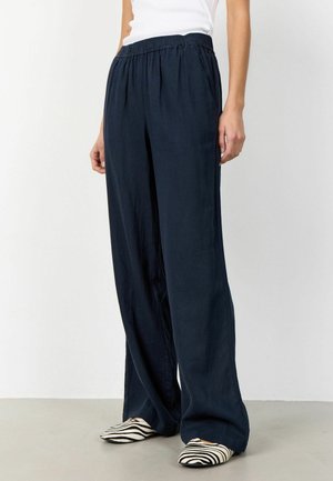 Žena stojaca na šedej podlahe, oblečená v širokých tmavomodrých nohaviciach s elastickým pásom a čierno-bielych pruhovaných špicatých balerínach.