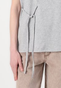 Gray cotton t-shirt with a knotted tie detail on the side, paired with beige textured trousers. Focus on fabric and design elements.