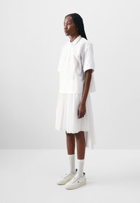 Hvit polo skjorte med krage og sidestriper, paret med en hvit plissert skjørt. Modellen har på seg hvite joggesko og ankelstrømper.