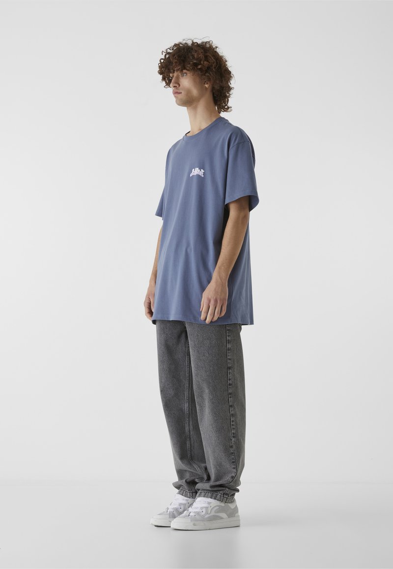 Nooruk, kellel on lokkis juuksed, seisab lahtises sinises T-särgis, hallides lahtistes teksapükstes ja valgetes tossudes, taustal on valge sein.