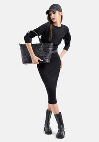 Borsa a tracolla nera trapuntata con logo circolare, indossata con una gonna nera aderente e un maglione nero, abbinata a stivali neri al ginocchio e un berretto.
