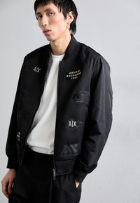 Giacca bomber nera realizzata in tessuto a rete, con toppe ricamate recanti i loghi "Armani Exchange" e "AIX". Chiusura con zip e polsini a coste.