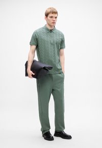 En ung man som bär en texturerad grön polo och matchande byxor, håller en stor svart vikbar läderclutch, stående mot en vit bakgrund.