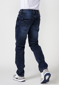 Blugi din denim de culoare albastră închisă, cu croială slim, având două buzunare pe spate și un tiv contrastant cusut. Sunt purtate adidași cu talpă din cauciuc.