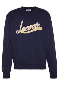 Mörkblå sweatshirt med rund hals, prydd med en stor gul "Lacoste"-logotyp över bröstet, ribbade ärmslut och nederkant, samt en liten grön logotyp på ärmen.