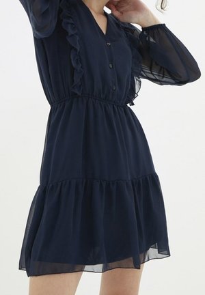 Kobieta ubrana w granatową, przezroczystą sukienkę z długim rękawem, z falbankami, guzikami z przodu i marszczonym w pasie, stoi z uniesioną jedną ręką.