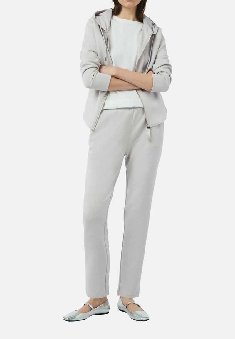 Jasnoszara bluza z kapturem i zamkiem z przodu, zestawiona z dopasowanymi zwężającymi się spodniami dresowymi oraz srebrnymi płaskimi butami. Gładka faktura materiału.