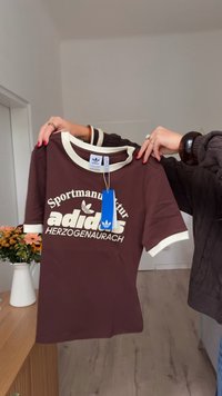 Hender holder en brun Adidas t-skjorte med hvit krage og mansjetter, med teksten "Sportmanufaktur Herzogenaurach" og et blått merkebånd festet.