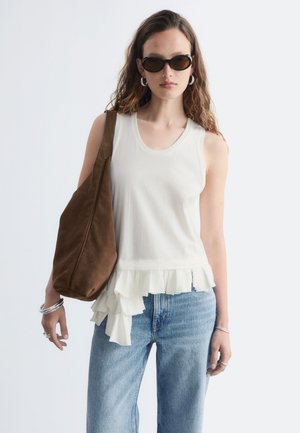 Femme aux cheveux bruns ondulés porte des lunettes de soleil noires, un top blanc sans manches à volants, un jean bleu clair, des bijoux en argent, et porte un grand sac à épaule marron.