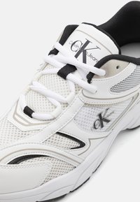 Baltas sporta apavi ar sietu un ādas virsu, kontrastējošiem melniem akcentiem, logs sānos, saisēšanas dizains, mīksts apkakle un teksturēta zole.