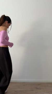 Žena s tmavými vlasmi v copíku si upravuje ružový top s odkrytými ramenami, stojí na drevenej podlahe pred jednoduchou bielou textúrovanou stenou.