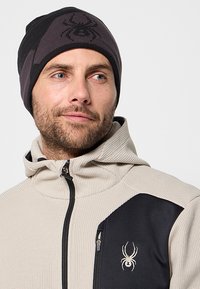 Berretto a maglia nero e grigio con un logo a ragno; indossato con una giacca beige testurizzata con un accento nero e una tasca con zip.