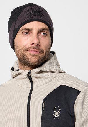 Berretto a maglia nero e grigio con un logo a ragno; indossato con una giacca beige testurizzata con un accento nero e una tasca con zip.