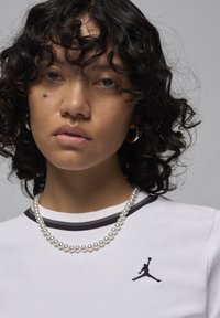 White short-sleeve shirt with black trim featuring a black Jordan logo; model wears a pearl necklace and has curly hair.