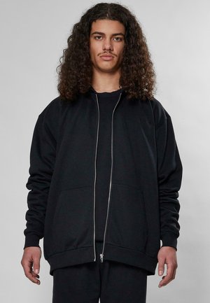 Svart zip-up hettegenser med lange ermer, ribbestrikkede mansjetter, to frontlommer og en avslappet passform. Tøyet har en glatt og myk tekstur.