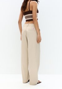 Kvinde iført beige bredbenede bukser og en ærmeløs brunstribet crop top, stående sidelæns på en hvid baggrund.