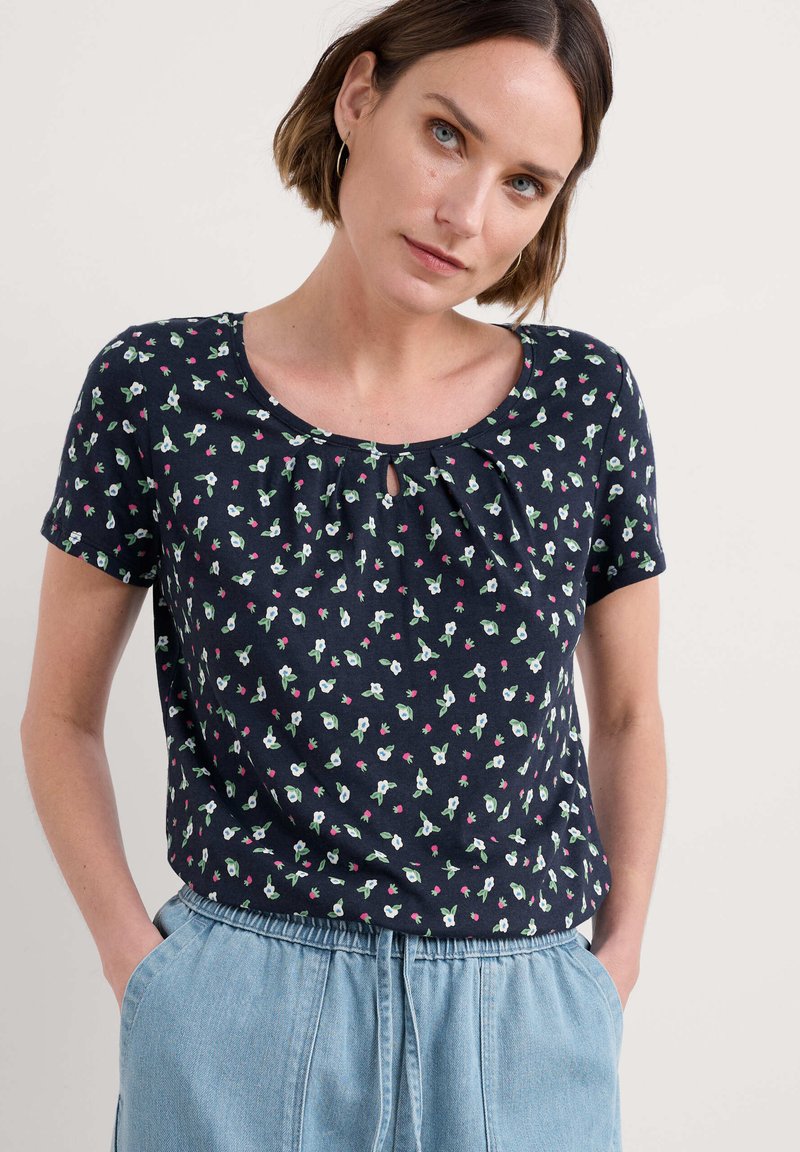 Jūrinės mėlynos trumpomis rankovėmis viršutinė dalis su gėlių raštu, apvaliu iškirpimu ir rakto angos detalėmis priekyje, pagaminta iš minkštos medžiagos.