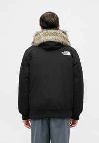 Jachetă puffer neagră cu guler de blană detașabil, cu manșete și tiv ribbate, și un logo alb pe umărul stâng.