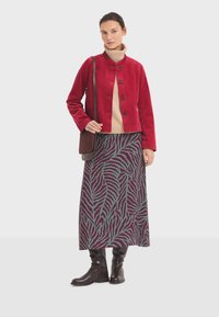 Rode corduroy jas met vijf knopen, beige coltrui eronder, en een gemusterde maxi-rok met een groen en burgundy bladdesign. Bruine laarzen tot de knie.