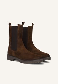 Een paar bruine suède Chelsea-boots met zwarte elastische zijpanelen en dikke rubberen zolen, schuin naar rechts geplaatst op een lichte achtergrond.