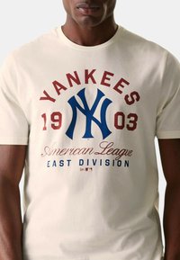 Camiseta de algodón crema de manga corta con el logo de los Yankees en rojo y azul, "1903" y "División Este de la Liga Americana" impresos en la parte frontal.