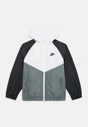 Chaqueta cortavientos con capucha blanca, mangas negras, parte inferior delantera gris, cremallera frontal, bolsillos con cremallera y logo negro de Nike en el pecho.