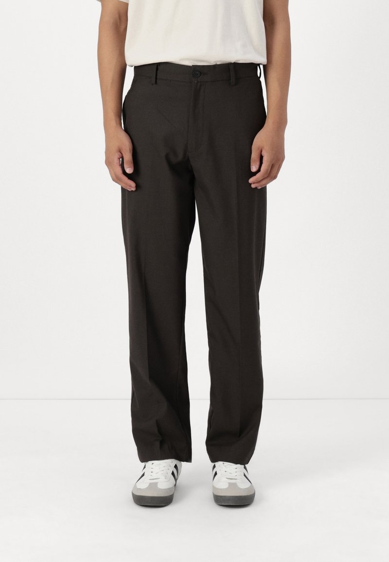Personne portant un pantalon noir droit et des baskets blanches avec des rayures noires, debout sur un fond blanc uni.
