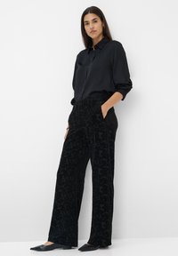Schwarze Bluse mit einem Knopfleiste-Design, kombiniert mit schwarzen, samtigen, weit geschnittenen Hosen mit einem dezenten floralen Muster. Schwarze spitze Ballerinas.