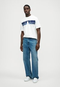 T-shirt branca com design gráfico azul e decote redondo, combinada com jeans azul claro de perna reta e ténis brancos. Roupa simples e casual.