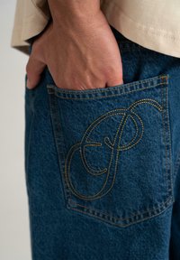 Jean en denim bleu avec un logo brodé distinctif sur la poche arrière. Présente une coupe standard et une finition en tissu texturé.