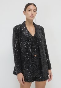 Donna in blazer di paillettes nere, pantaloni corti di paillettes coordinati e orecchini a catena lunghi, in piedi contro uno sfondo chiaro e uniforme.