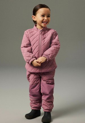 Smilende barn stående iført quiltet lyserød jakke og bukser med sorte støvler mod en ensfarvet grå baggrund.