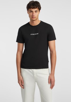 Svart bomull T-skjorte med korte ermer, med en hvit "GUESS JEANS" logo sentrert på brystet. Modellen kombinerer den med hvite bukser.