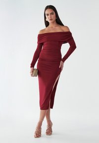 Burgundská šaty bez ramienok s nahrnutým detailom, priliehavým strihom, rozparkom po stehná a dlhými rukávmi. Spárované so sandálmi so zlatými akcentmi.