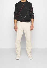 Černý svetr s barevnými diagonálními pruhy, spárovaný s krémovými kalhotami a bílými teniskami. Uvolněný střih s žebrovanými manžetami.