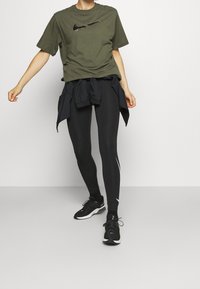 Camiseta de algodón verde oliva con el logo negro de Nike, combinada con leggings negros que tienen acentos laterales blancos y zapatillas deportivas negras.