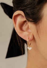 Orecchino a cerchio dorato con un ciondolo a forma di cuore. Il cuore presenta piccoli strass trasparenti, creando un accento scintillante. Texture liscia.