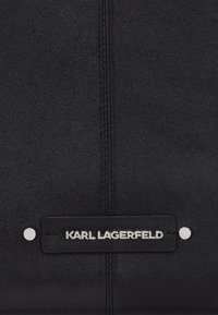Sac à main en cuir noir avec une surface texturée, comportant une étiquette logo en métal argenté et des détails de couture visibles.