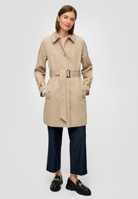 Beige trenchcoat met een ceintuur in de taille, knoopsluitingen en twee voorkussens. Gestyled met donkerblauwe wijde broek en zwarte instappers.