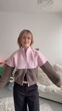 Joven sonriente con los brazos extendidos, vistiendo una chaqueta de forro polar con cremallera en rosa y marrón en una sala de estar acogedora.