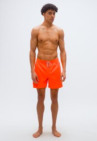 Νεαρός άνδρας με σγουρά μαλλιά, όρθιος και ξυπόλητος, φοράει φωτεινά πορτοκαλί μαγιό μπροστά σε λευκό απλό φόντο.