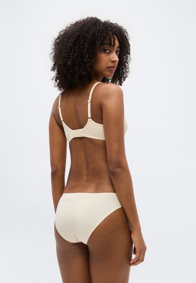 Γυναίκα με κατσαρά μαλλιά που φοράει μπεζ σουτιέν και αντίστοιχο εσώρουχο, στέκεται με την πλάτη προς την κάμερα και κοιτάζει πάνω από τον ώμο.