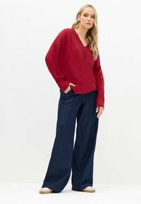 Roter, locker sitzender V-Ausschnitt-Pullover mit gerippten Bündchen, kombiniert mit weit geschnittenen, dunklen Denim-Jeans und hellen Slippern. Minimalistischer Hintergrund.