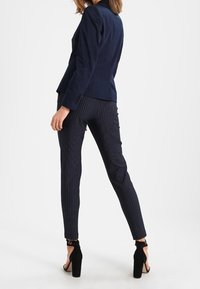 Granatowa marynarka o dopasowanym kroju, zestawiona z granatowymi spodniami w prążki. Czarne sandały na wysokim obcasie z odkrytymi palcami, widoczny pasek wokół kostki.