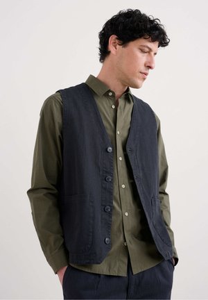 Chaleco de algodón negro con cinco botones y dos bolsillos, usado sobre una camisa de cuadros verde olivo con cuello clásico y estilo de botón.