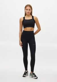 Zwarte sportbeha met brede banden en logodetail, gecombineerd met hoge taille zwarte legging. Model draagt atletische schoenen met kleuraccenten.