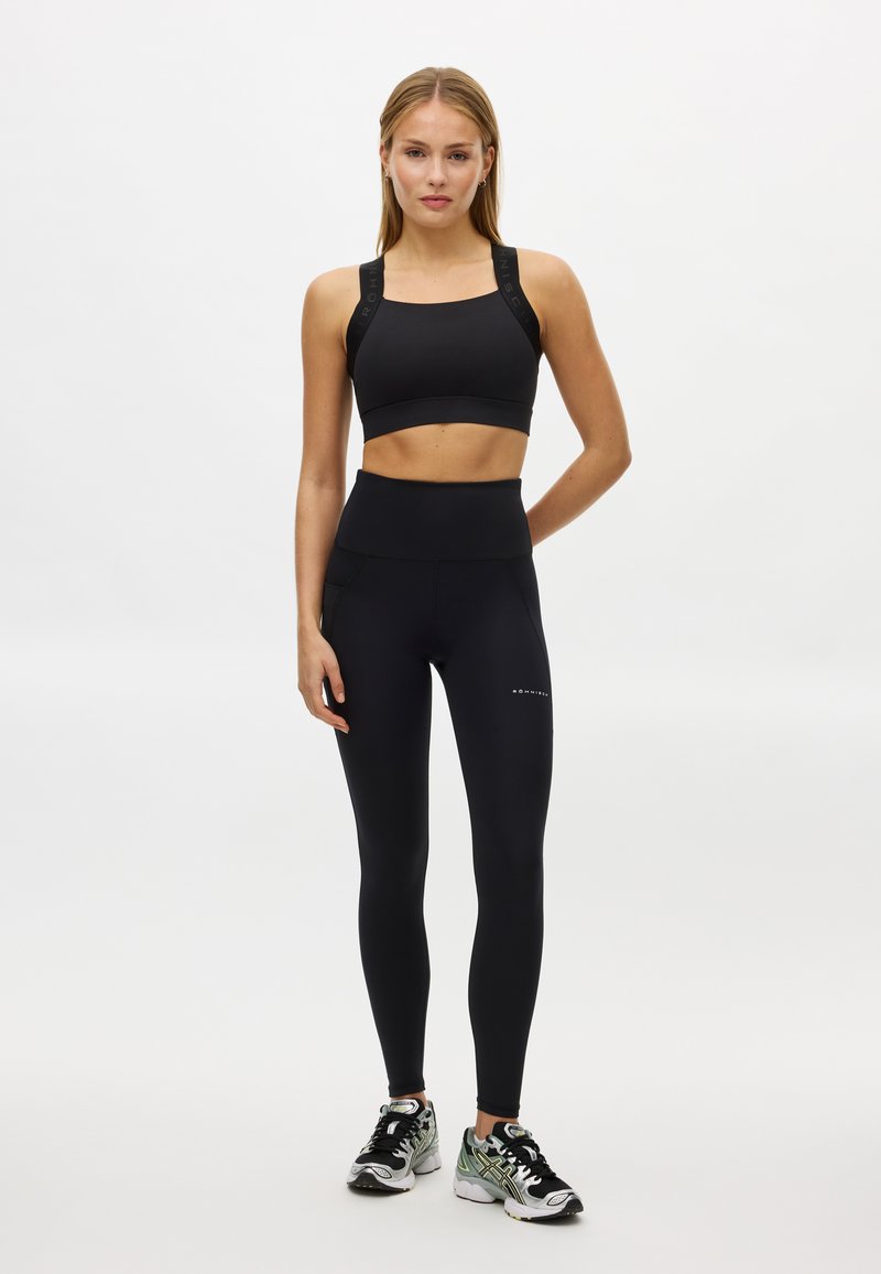 Zwarte sportbeha met brede banden en logodetail, gecombineerd met hoge taille zwarte legging. Model draagt atletische schoenen met kleuraccenten.