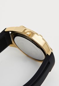 Montre-bracelet en métal doré avec un bracelet en caoutchouc noir, vue de côté montrant la couronne et le détail texturé du bracelet sur fond blanc.